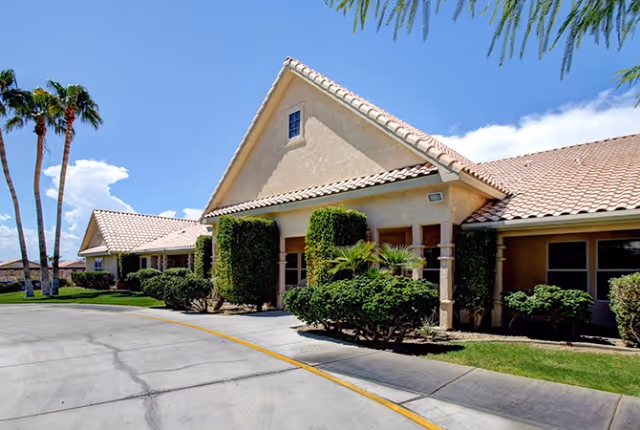 Exterior view of a single-story senior living facility building with beige stucco walls and a tiled roof. The building is surrounded by neatly trimmed bushes and palm trees under a clear blue sky.