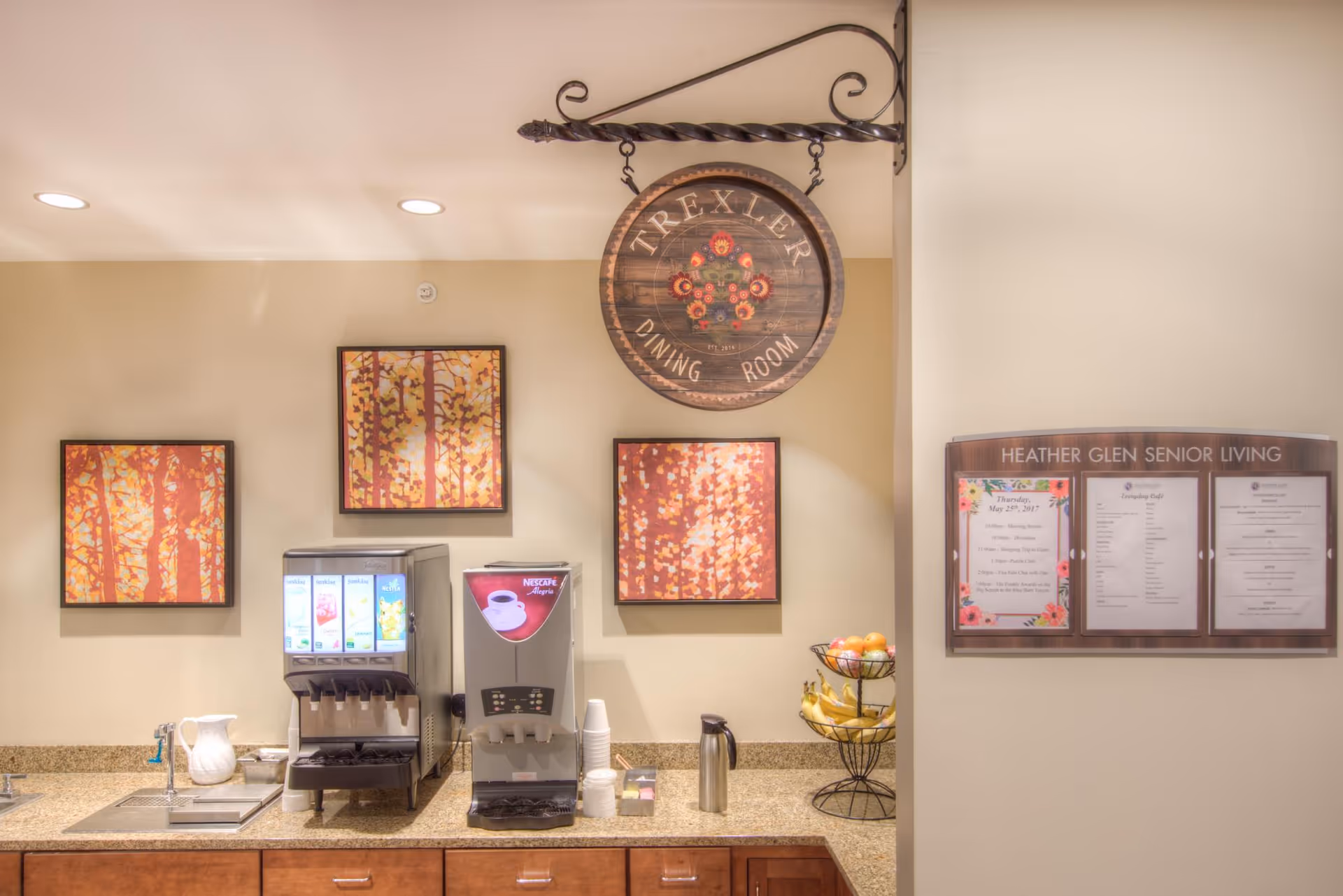 Counter with beverage dispensers, a fruit stand, and framed artwork beneath a hanging 'Trexler Dining Room' sign and a 'Heather Glen Senior Living' bulletin board.
