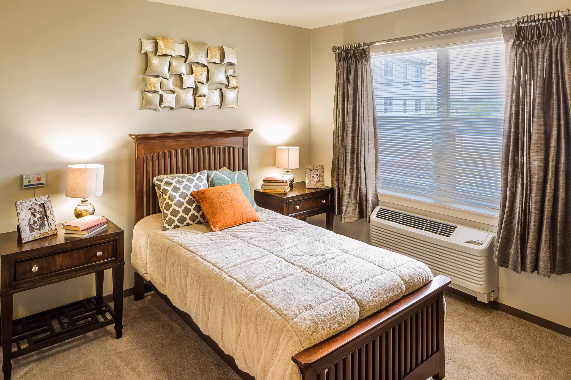 A cozy bedroom with a wooden bed frame and a beige quilted bedspread. The bed is adorned with three decorative pillows in orange, teal, and patterned gray. On either side of the bed are matching wooden nightstands with table lamps, books, and framed photos. A large window with brown curtains and blinds lets in natural light, and a wall-mounted air conditioning unit is below the window. A decorative metal wall art piece hangs above the bed.