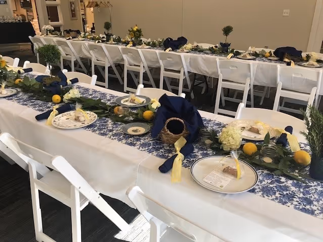 Long dining tables set up with white tablecloths and blue floral runners, decorated with small plants, lemons, and napkins folded with yellow tassels. White folding chairs are arranged along both sides of the tables in a well-lit room.