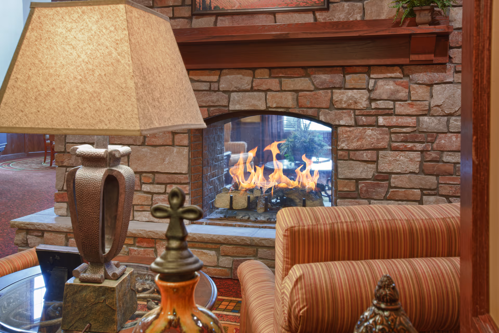Cozy interior seating area with a striped upholstered armchair, a stone fireplace with a burning fire, a decorative table lamp, and ornamental objects on a glass-top table.
