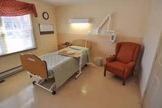 A simple patient bedroom with a hospital bed, overbed table, red armchair, wall-mounted light and a window with blinds.