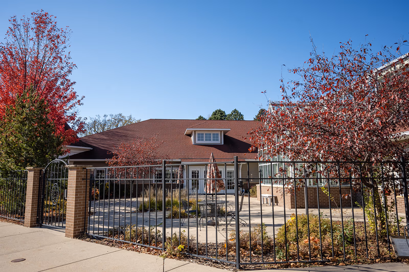 Front exterior of a single-story cottage with a fenced patio, red roof, and autumn trees.