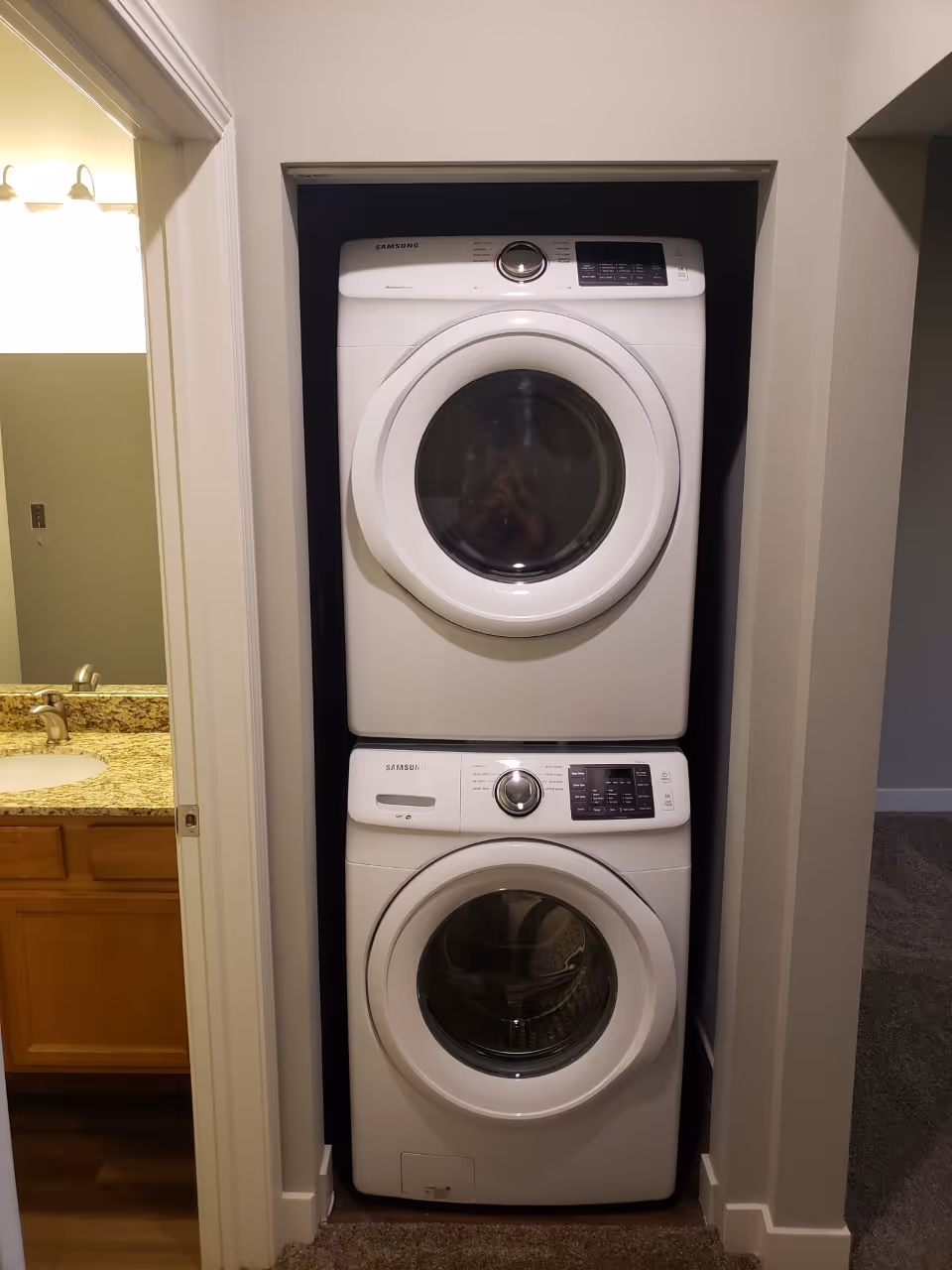 Stacked white Samsung washer and dryer units installed in a recessed wall space with a bathroom visible to the left and carpeted flooring in front.
