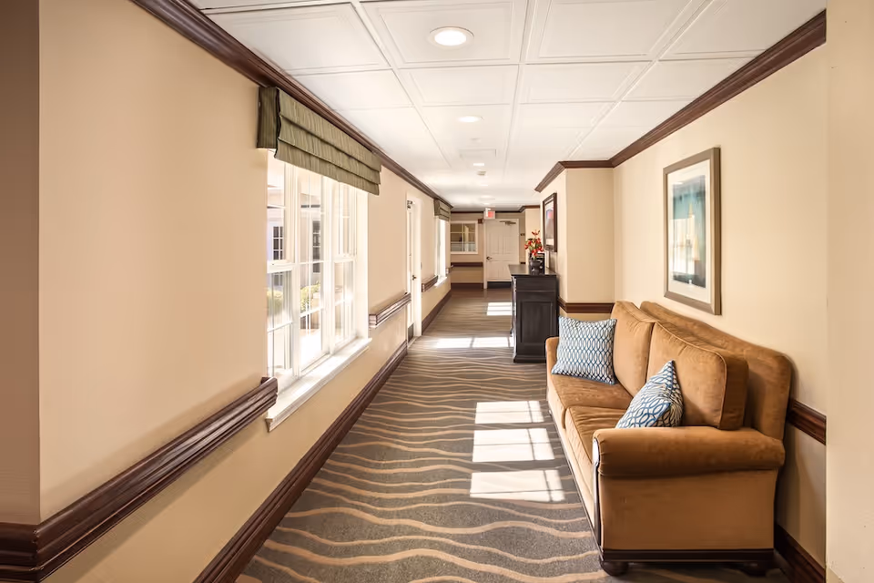 A well-lit hallway in a senior living facility with beige walls, brown trim, and a patterned carpet. On the right side, there is a brown sofa with two patterned pillows and a framed artwork above it. On the left side, large windows with green valances allow natural light to enter. At the end of the hallway, there is a black cabinet with a flower arrangement on top.