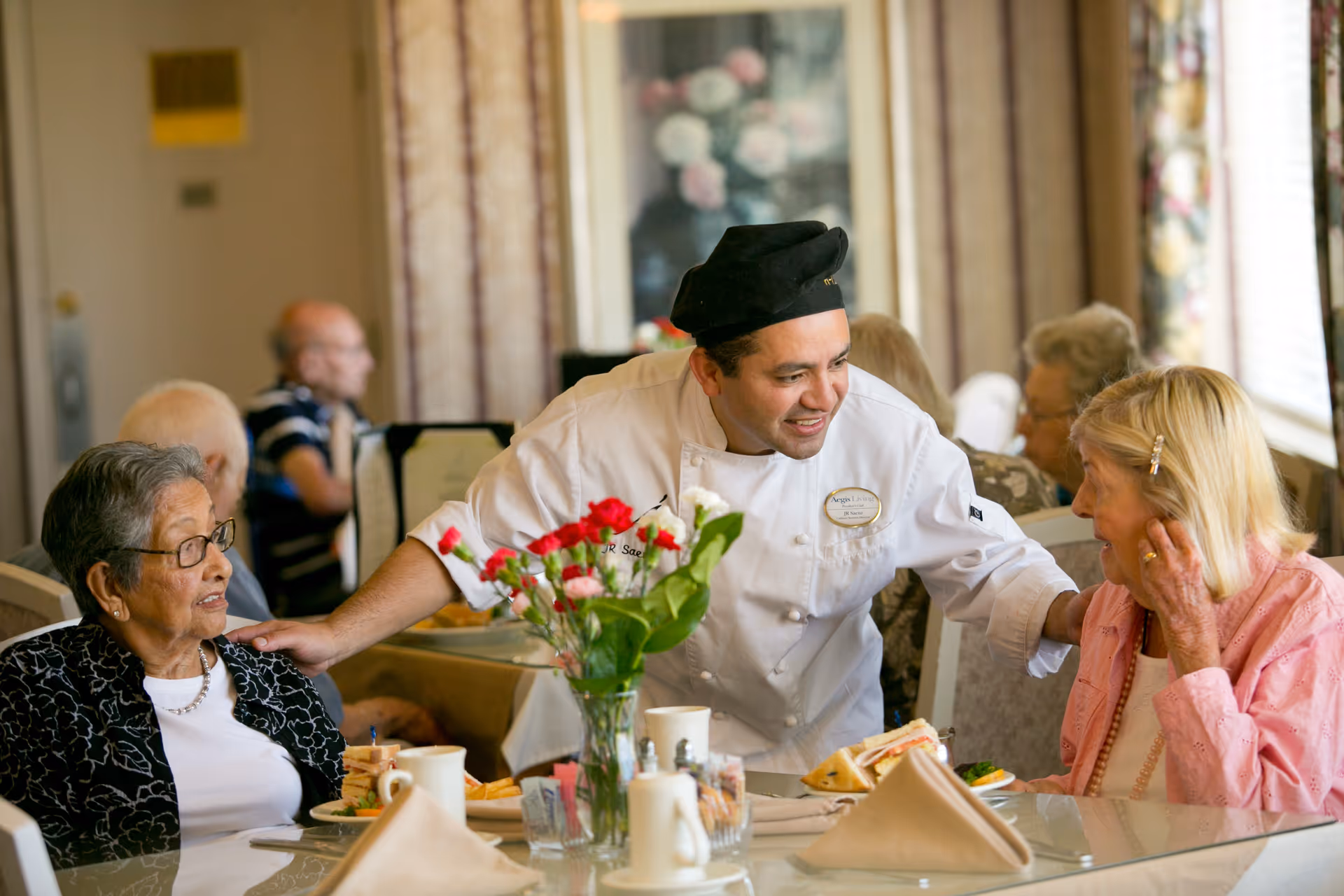 A chef in a white uniform and black hat is smiling and interacting warmly with two elderly women seated at a dining table in a well-lit room. The table is set with plates of food, cups, and a vase with red and white flowers. Other elderly people are visible in the background, seated and engaged in conversation.