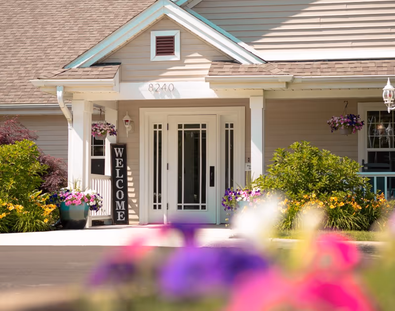 Front entrance of a building with a white door featuring glass panels, a 'WELCOME' sign hanging beside the door, and colorful flowers and greenery around the entrance. The building has beige siding and a brown shingled roof with the number 8240 above the door.