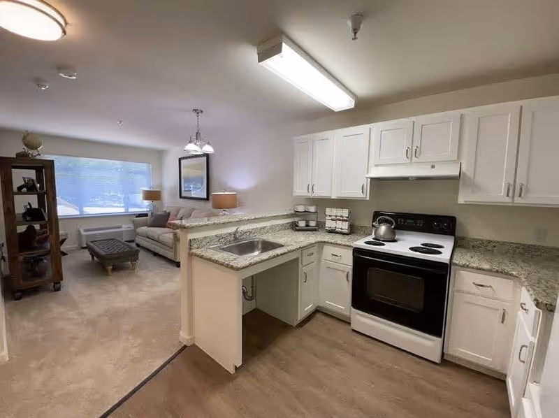 A bright and clean kitchen area with white cabinets, a granite countertop, a stainless steel sink, and a white electric stove with a kettle on it. The kitchen opens into a living room with a large window, a beige sofa, a cushioned ottoman, a wooden shelving unit with decorative items, and two table lamps.