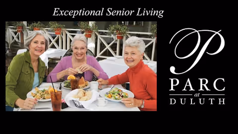 Three elderly women sitting at an outdoor dining table, smiling and enjoying a meal together with plates of food and drinks in front of them.