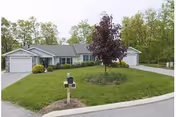 Single-story residential building with attached garages on both sides, a well-maintained lawn with a small tree and mailbox in front, surrounded by trees in the background.