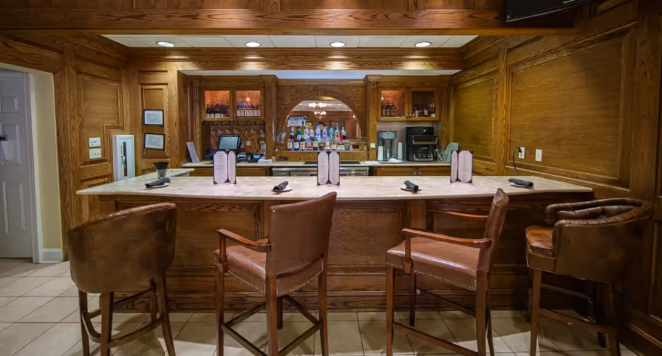 A cozy bar area with a marble countertop and four brown leather bar stools. Behind the bar, there are shelves stocked with various bottles of liquor, a coffee machine, and a cash register. The walls and cabinetry are made of warm wood paneling, and the floor is tiled.