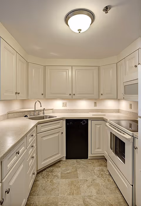 A clean, well-lit kitchen with white cabinetry, a stainless steel sink, a black dishwasher, a white electric stove with oven, a microwave, and beige tiled flooring.