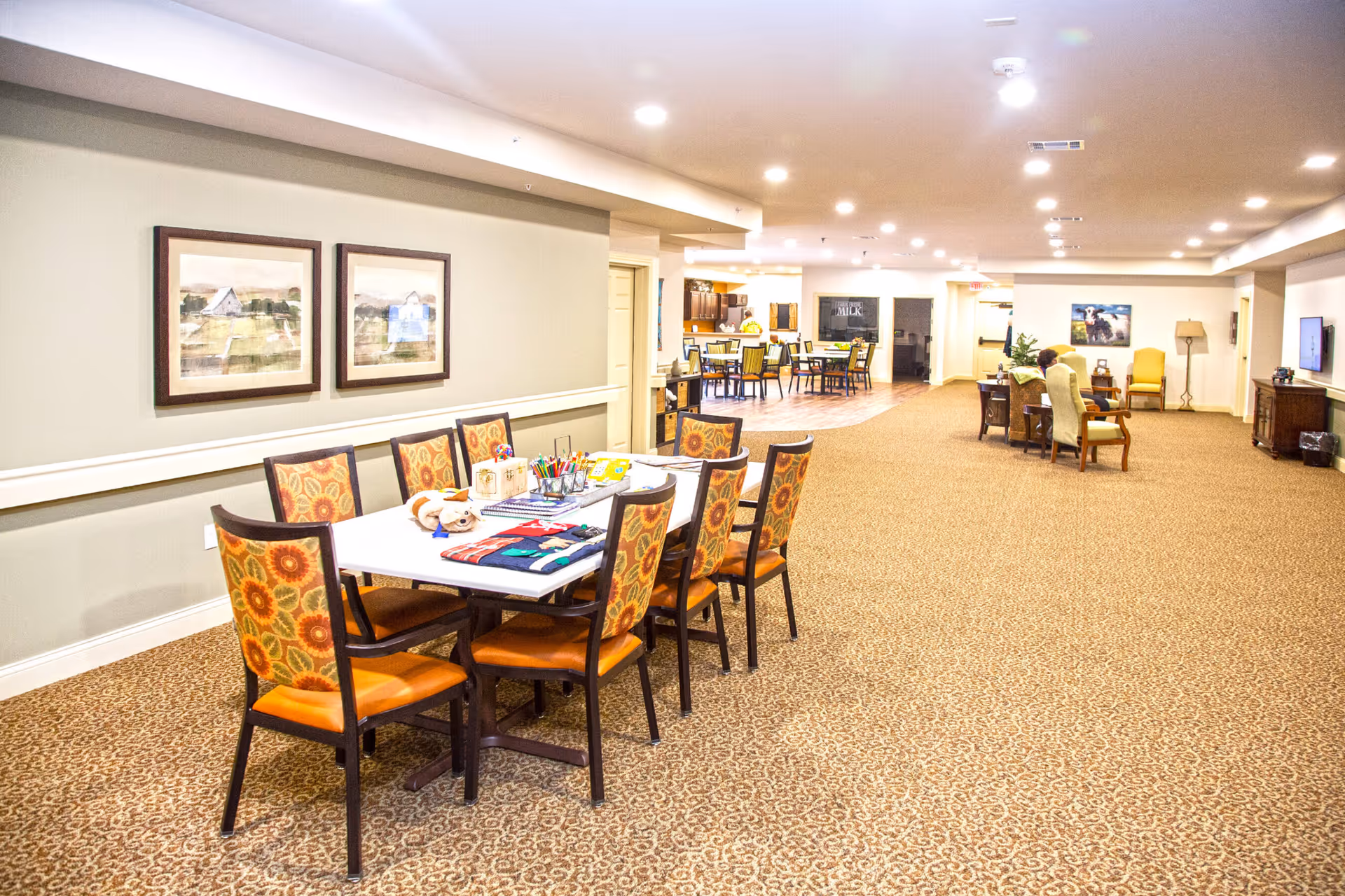 A spacious, well-lit common area in a senior living facility with a long table and eight chairs featuring floral upholstery. The table has various craft supplies and a stuffed animal on it. In the background, there are additional seating areas with chairs and tables, a television mounted on the wall, and framed artwork. The carpet is patterned and the walls are painted a light beige color.
