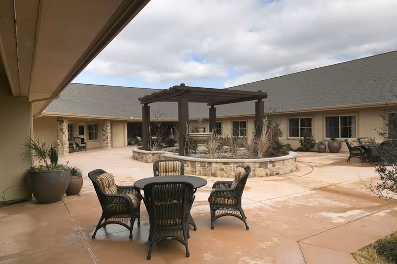 Outdoor courtyard area of a senior living facility with a round table and four wicker chairs with cushions. The courtyard features a stone planter with a wooden pergola and various plants. The building surrounds the courtyard with multiple windows and doors visible under a cloudy sky.