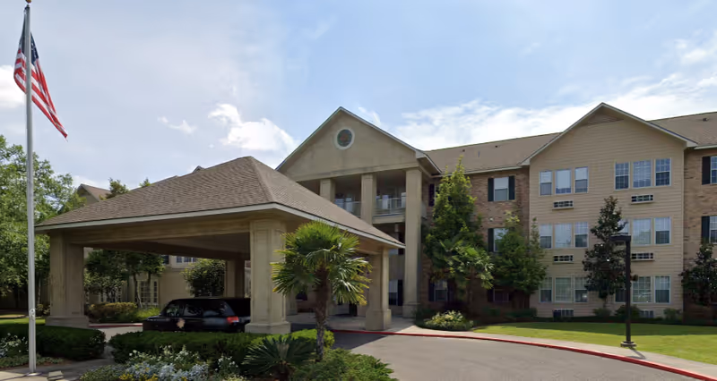 Exterior view of a senior living facility with a covered driveway entrance, an American flag on a flagpole, and a three-story building with multiple windows surrounded by trees and landscaping under a partly cloudy sky.