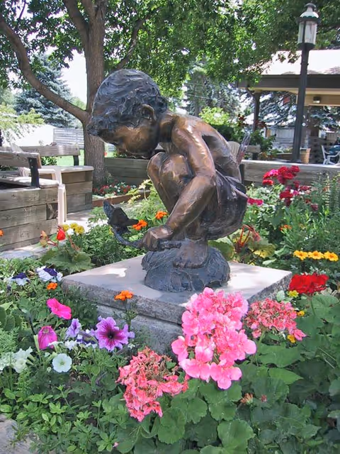 Bronze statue of a crouching child holding a bird on a pedestal surrounded by colorful flowers and garden benches.