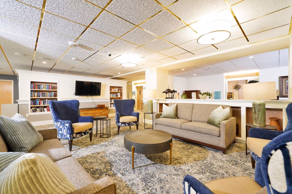 A bright and spacious living room area in a senior living facility featuring two beige sofas with green pillows, four blue patterned armchairs, a round coffee table in the center, a patterned area rug, built-in bookshelves filled with books, a wall-mounted flat-screen TV, and modern ceiling lights.
