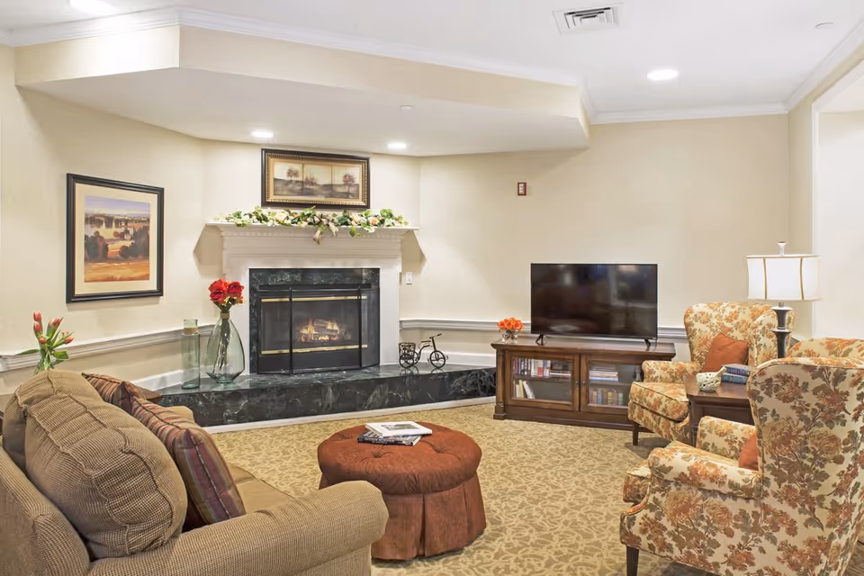 A cozy living room with a brown sofa, two floral armchairs, a round brown ottoman, a wooden TV stand with a flat-screen TV, and a fireplace decorated with a floral garland and framed artwork above it. The room has beige walls and carpet with a floral pattern.