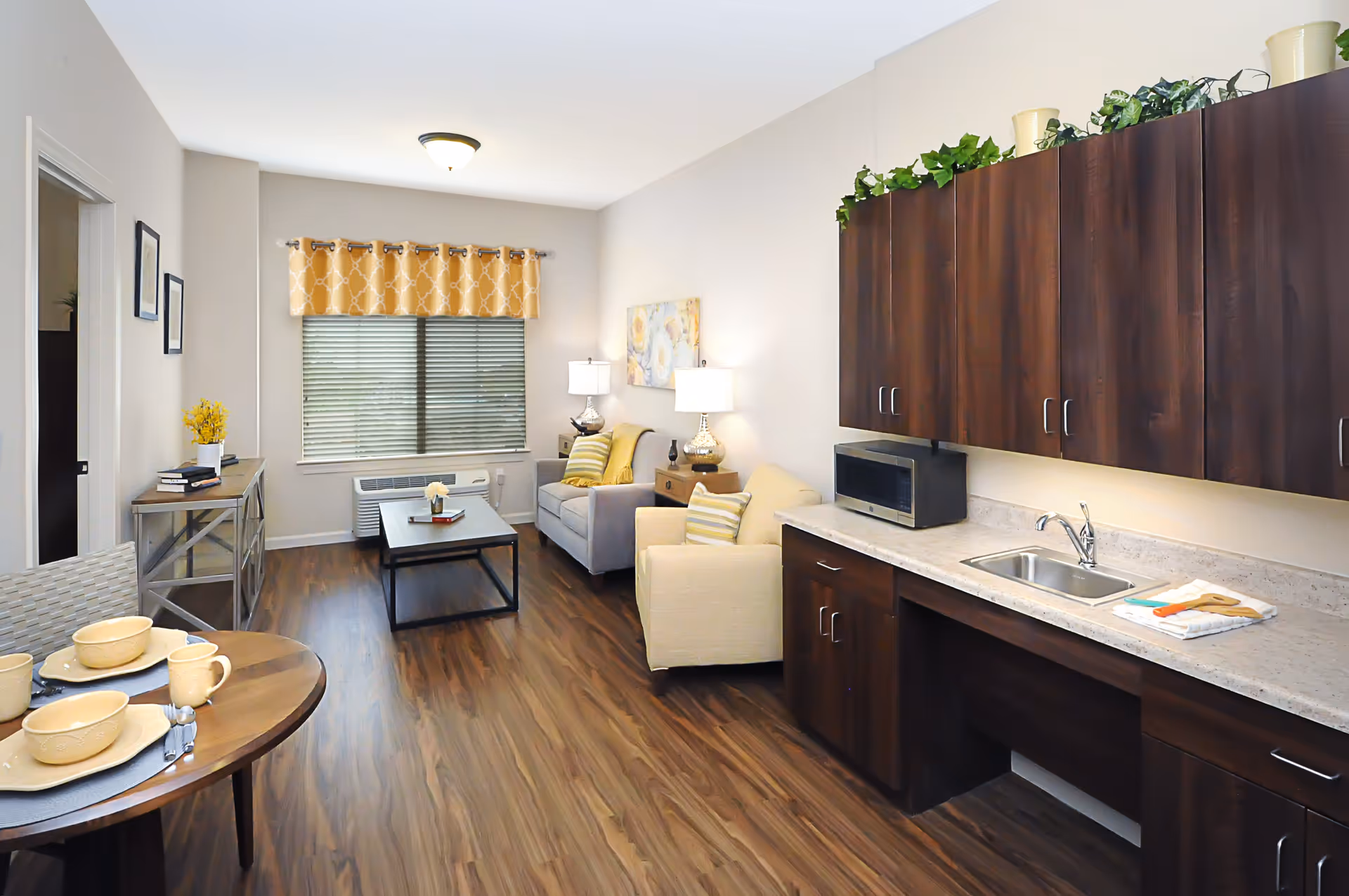 A bright and cozy senior living apartment featuring a small kitchen area with dark wooden cabinets, a countertop with a sink, and a microwave. The living area has a beige armchair, a gray loveseat with striped pillows, two table lamps, a coffee table, and a window with yellow patterned curtains. A round dining table with beige dishware is visible in the foreground, and the floor is covered with wood-style laminate.