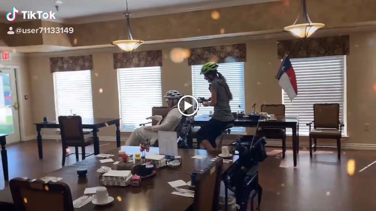 A senior woman and a younger person wearing helmets are seated in a spacious room with large windows covered by blinds. The room has several tables and chairs arranged around, with various items like cups, tissues, and papers on the tables. A flag is visible near the windows, and the floor is wooden.