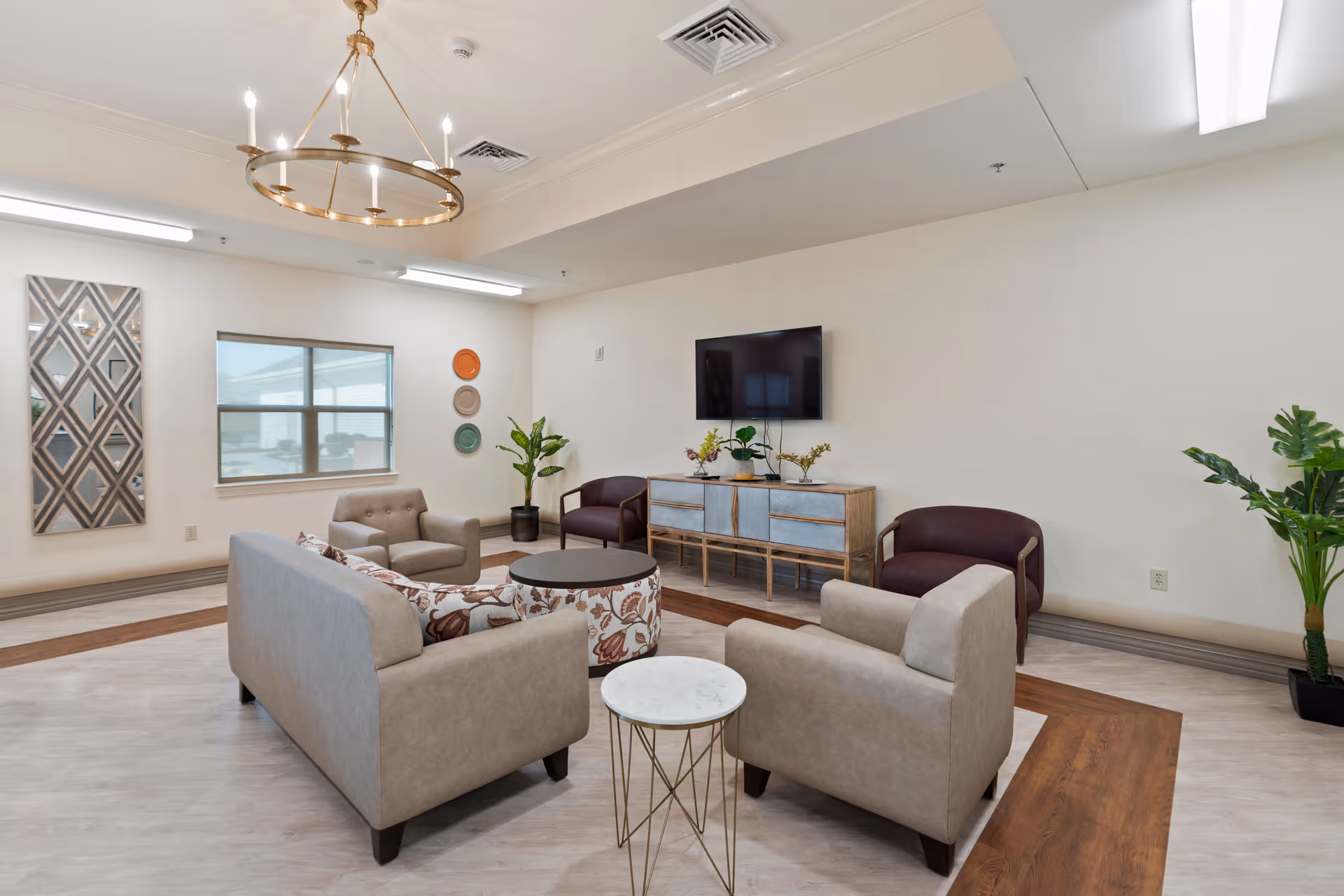 A bright and modern living room area with beige and patterned sofas, two maroon armchairs, a round coffee table, and a small round side table. A wooden console with plants and a flat-screen TV mounted on the wall is visible. The room has a large window, decorative wall art, potted plants, and a circular chandelier hanging from the ceiling.