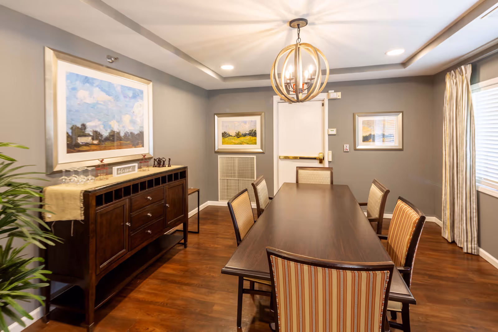 Well-lit dining room with a long wooden table surrounded by upholstered chairs, a sideboard, framed artwork, and a chandelier.
