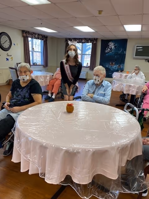 A group of elderly people sitting around tables covered with plastic tablecloths in a nursing home common area. A young woman wearing a crown, sash, and face mask stands behind one of the tables. The room has windows with curtains, a clock on the wall, and a Halloween-themed decoration in the background.