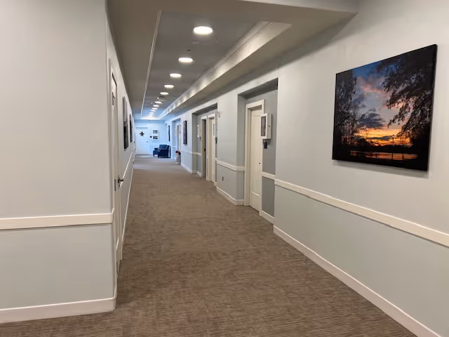 A long, well-lit hallway in a senior living facility with beige carpet and light gray walls. Several doors line the hallway on both sides, and framed pictures hang on the walls. At the far end, there is a seating area with blue chairs and a small table.