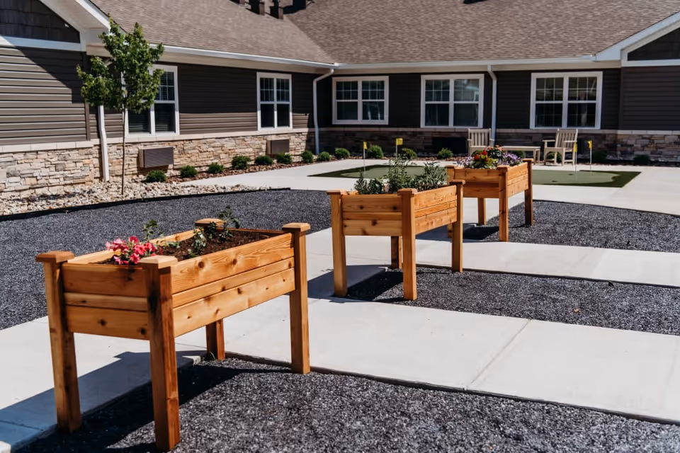 Outdoor courtyard area at Shelby Farms Senior Living featuring three wooden raised garden beds with plants and flowers, surrounded by gravel and concrete walkways. The building exterior has stone and siding with multiple windows and a small tree nearby.