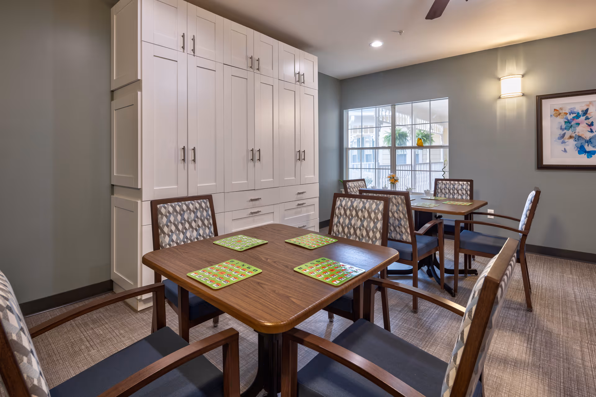 Dining area with wooden tables, patterned chairs, green placemats and tall white storage cabinets by a window.
