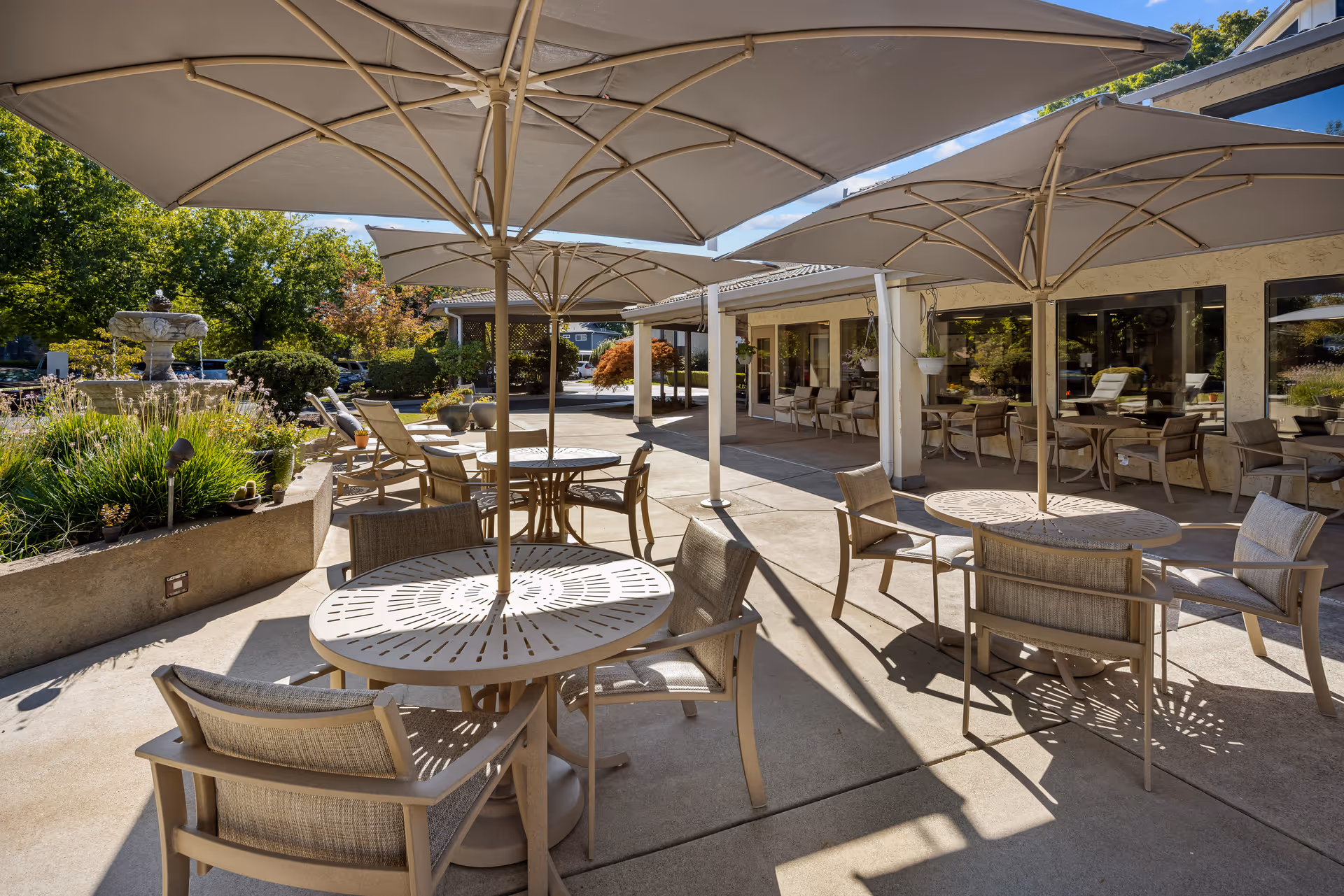 Outdoor patio area at Provincial Chico Senior Living with round tables and chairs under large beige umbrellas, surrounded by greenery and a water fountain in the background.