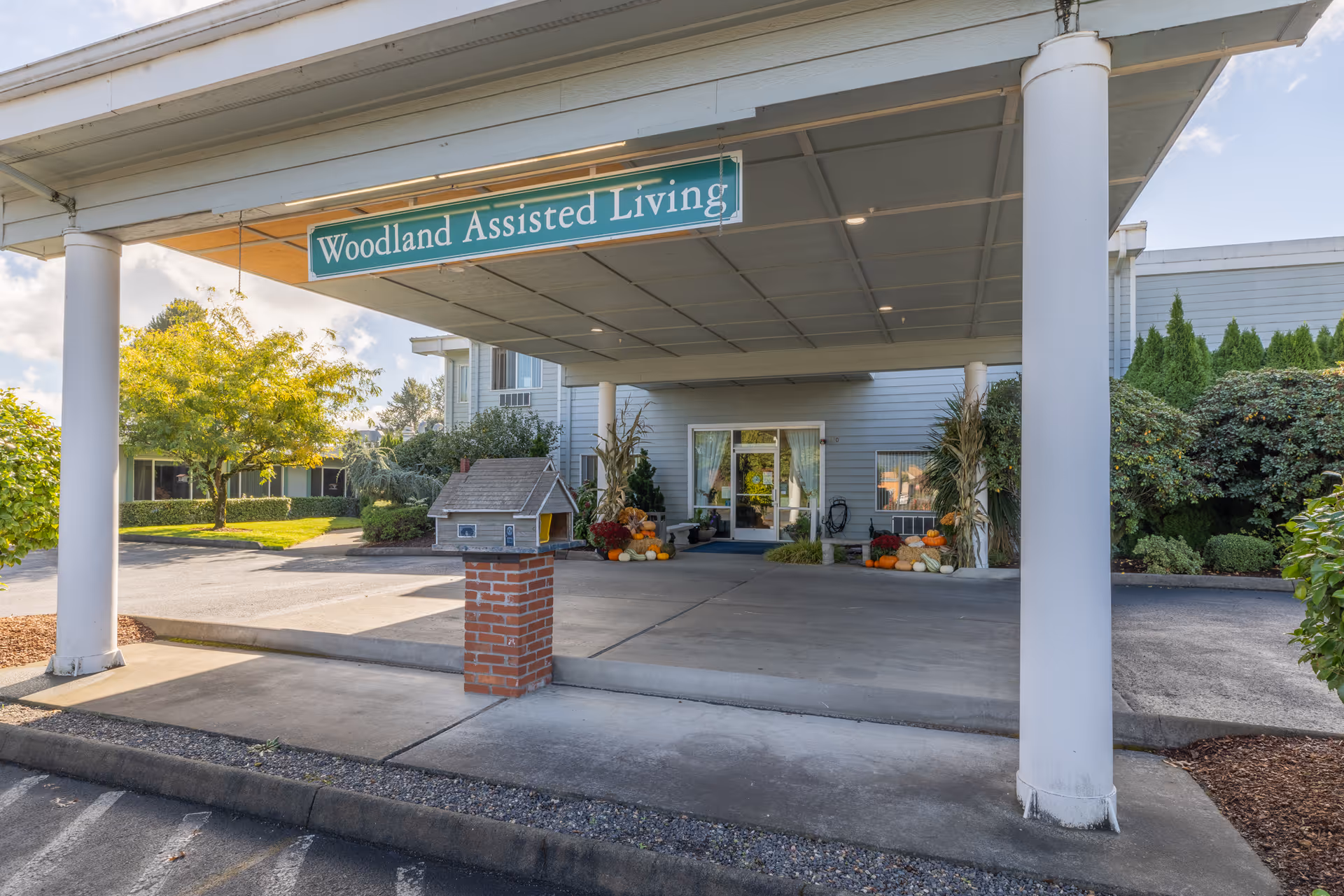 Covered entrance and drop-off canopy of Woodland Assisted Living with a hanging sign, white pillars, and seasonal pumpkins by the doorway.
