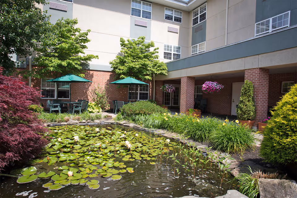 Outdoor courtyard area at Brandywine The Gables by Monarch featuring a pond with lily pads and blooming flowers, surrounded by lush greenery and trees. There are green patio umbrellas shading tables and chairs near the building with brick and beige walls, and hanging flower baskets add color to the covered walkway.