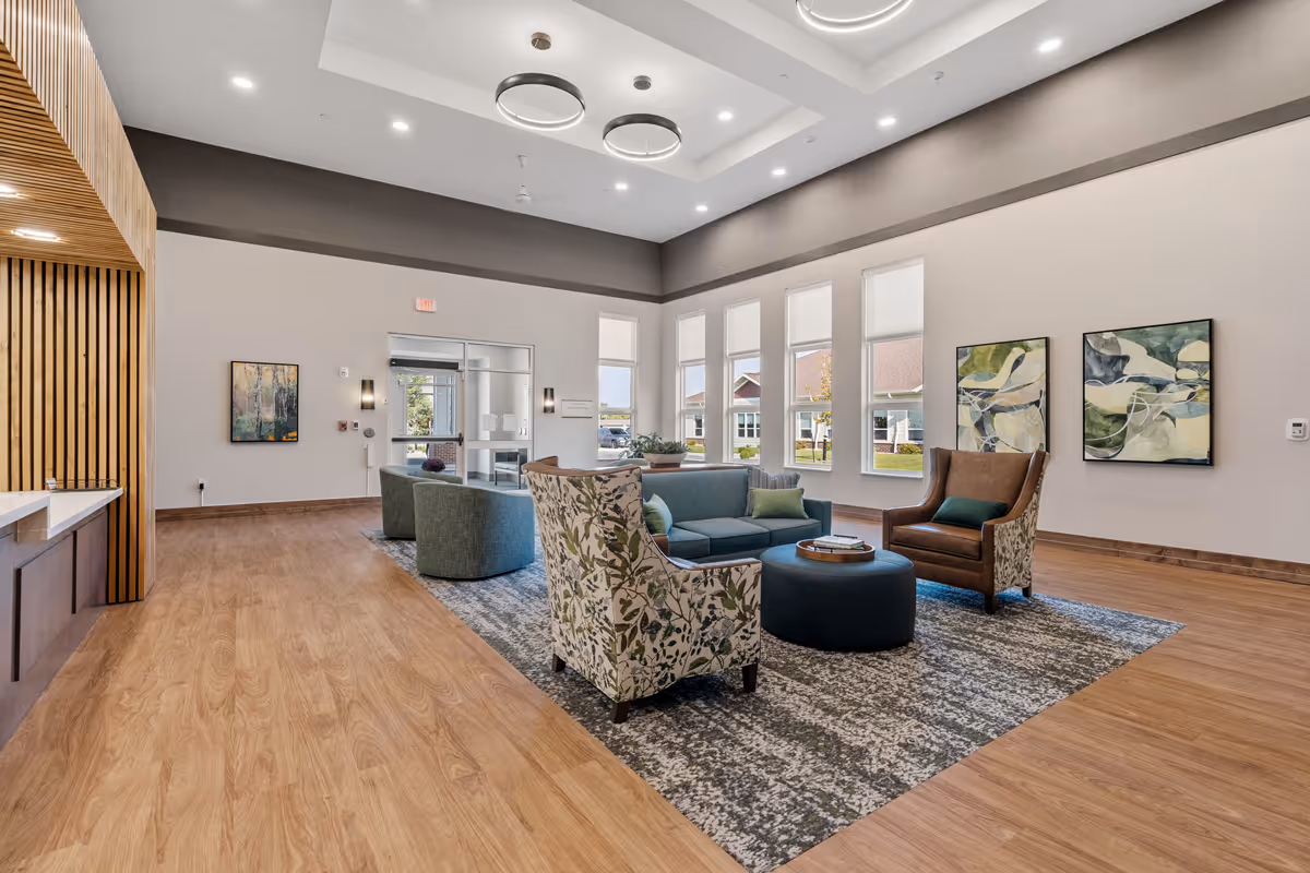 A spacious and modern living room area in a senior living facility with a high ceiling featuring recessed lighting and circular hanging lights. The room has large windows letting in natural light, a seating arrangement with a blue sofa, two patterned armchairs, a brown leather chair, and a round ottoman on a patterned rug. The walls are decorated with abstract paintings, and the floor is wood with a light finish.