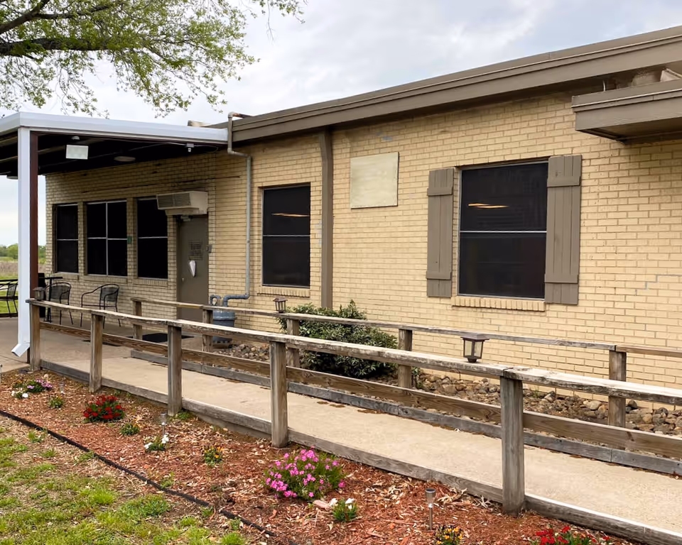 Exterior view of a single-story brick building with a covered patio area featuring black metal chairs. There is a concrete walkway with a wooden handrail leading to a door. The building has several windows with brown shutters, and there are small flower beds with colorful flowers along the walkway. A tree branch extends over the top left corner of the image.