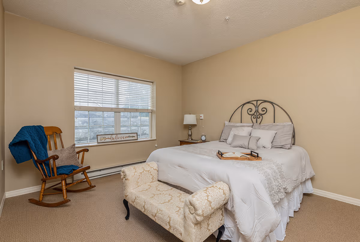 Cozy, neutral-toned bedroom featuring a neatly made bed with a decorative metal headboard, an upholstered bench at the foot, a rocking chair by the window, and a nightstand with a lamp.
