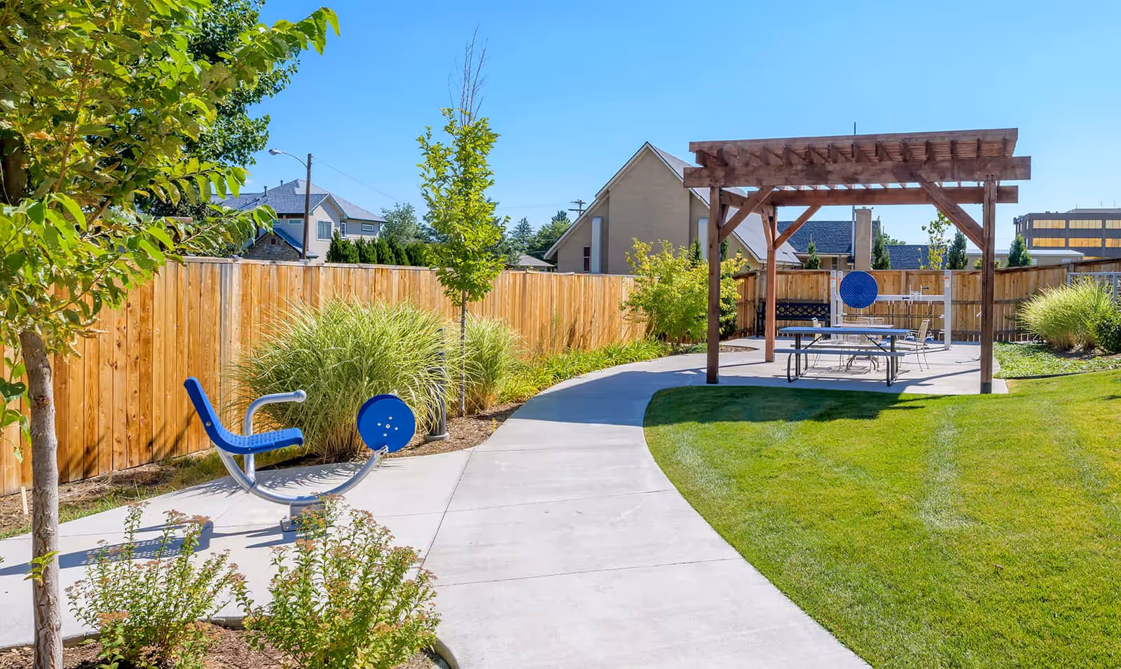 Outdoor area with a paved walkway, green grass, and a wooden pergola covering a picnic table and chairs. There is also a blue exercise machine and various trees and shrubs along a wooden fence under a clear blue sky.