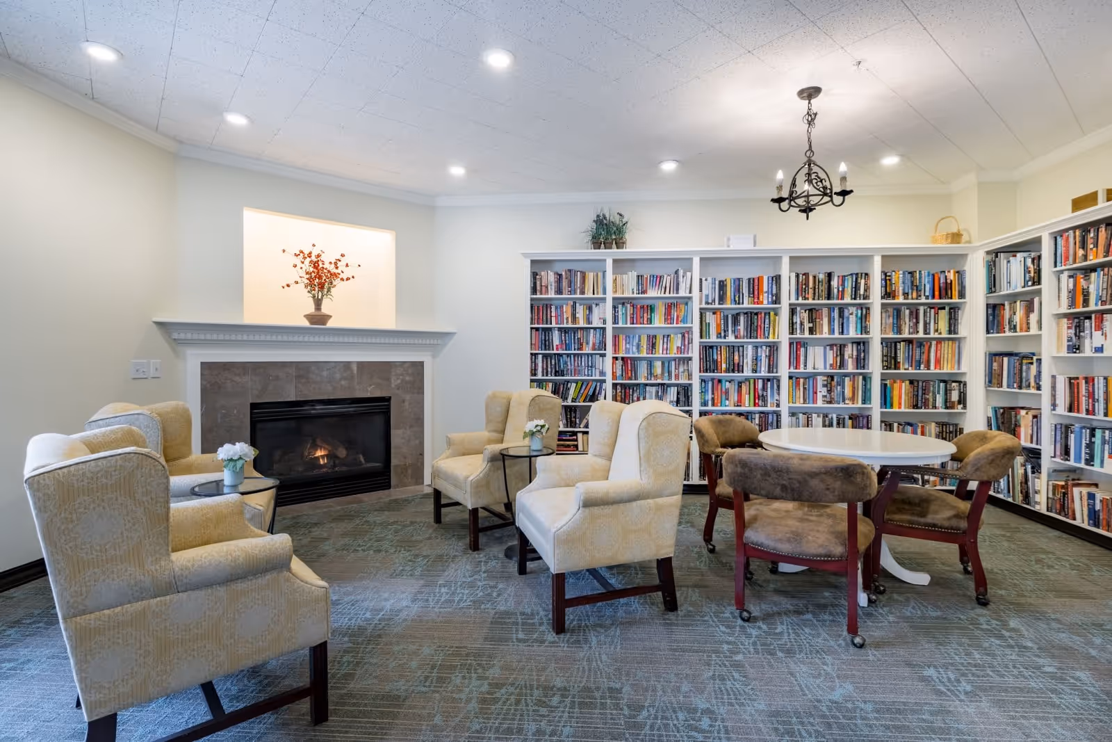 Cozy senior living library/lounge with bookshelves, upholstered armchairs, a fireplace and a round table.
