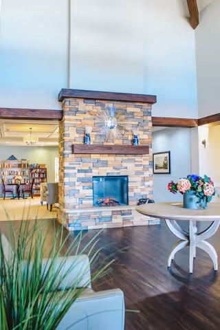 Interior view of a senior living facility common area featuring a stone fireplace with a decorative sunburst mirror above it. To the right is a round wooden table with a vase of colorful flowers. In the foreground, there is a green plant and part of a light-colored armchair. In the background, there is a room with bookshelves and seating.
