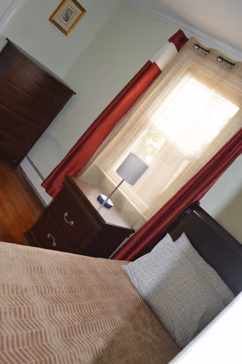 A bedroom with a bed covered in a beige blanket and a white pillow. Next to the bed is a wooden nightstand with a small lamp. A window with sheer beige curtains and red drapes is behind the nightstand. A wooden dresser and a framed picture are visible on the adjacent wall.