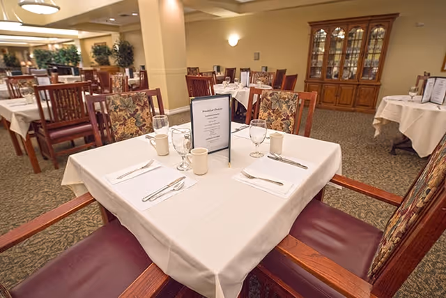 A dining room in a senior living facility with tables covered in white tablecloths, set with napkins, silverware, glasses, and coffee mugs. The chairs have wooden frames with patterned upholstery. In the background, there is a wooden china cabinet and more tables and chairs arranged neatly.