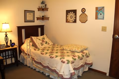 A small bedroom with a single bed covered in a patterned quilt and pillow. There is a wooden headboard, a nightstand with a lamp and a clock, and several decorative wall hangings. The room has beige walls and a carpeted floor.
