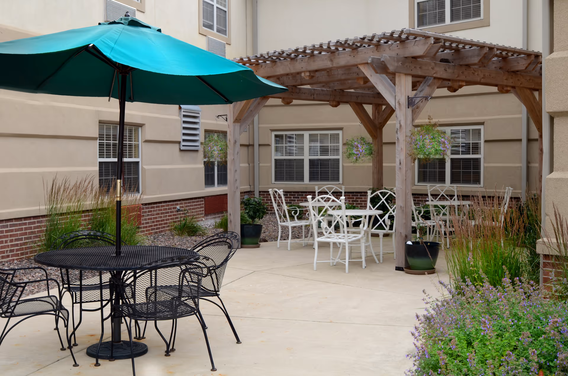 Outdoor patio area at Spring Mill Health Campus with black metal table and chairs under a green umbrella, white metal chairs and tables under a wooden pergola, potted plants, and surrounding building walls with windows.