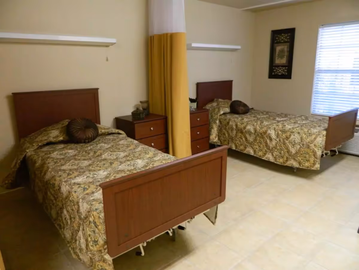 A nursing facility bedroom with two single beds separated by a yellow privacy curtain. Each bed has a wooden headboard and footboard, patterned bedspreads, and a round decorative pillow. Between the beds are two wooden nightstands with drawers and decorative items on top. A window with blinds is on the right wall, and a framed picture hangs on the wall next to the window.