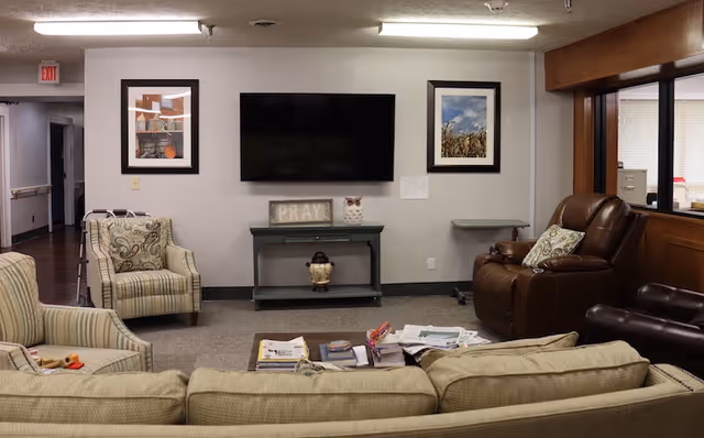 A cozy living room area in a senior care facility with a beige couch, two armchairs, and a brown recliner. A flat-screen TV is mounted on the wall above a small green table with decorative items including a sign that says 'PRAY' and an owl figurine. Two framed pictures hang on the wall on either side of the TV. A coffee table in front of the couch holds magazines and newspapers. The room has neutral-colored walls and carpeted flooring.