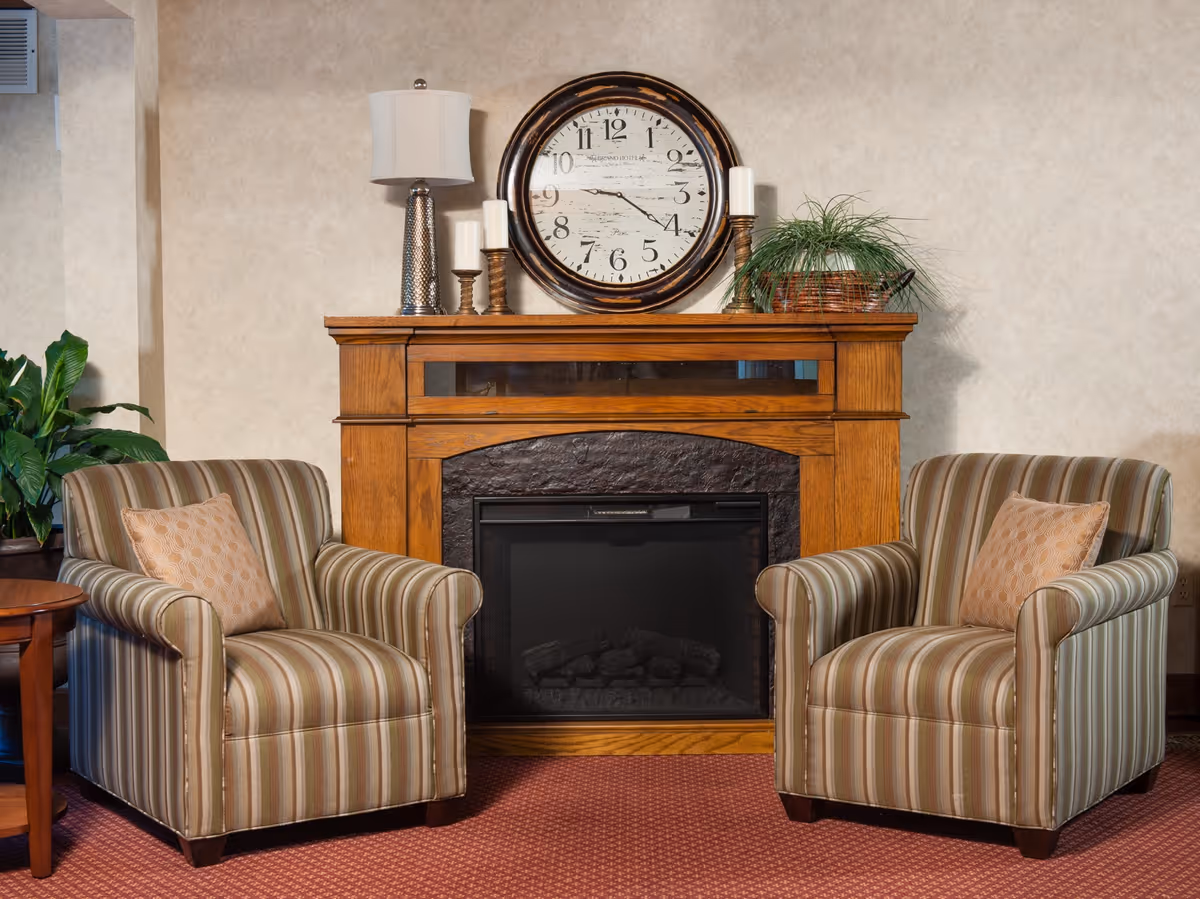 Two striped armchairs flanking a wooden fireplace mantel topped with a large round clock, lamp, candles, and a potted plant.
