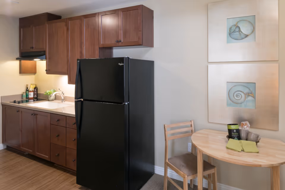 Kitchenette with dark wood cabinets, a black refrigerator, a sink and a small round dining table with a chair beneath framed wall art.
