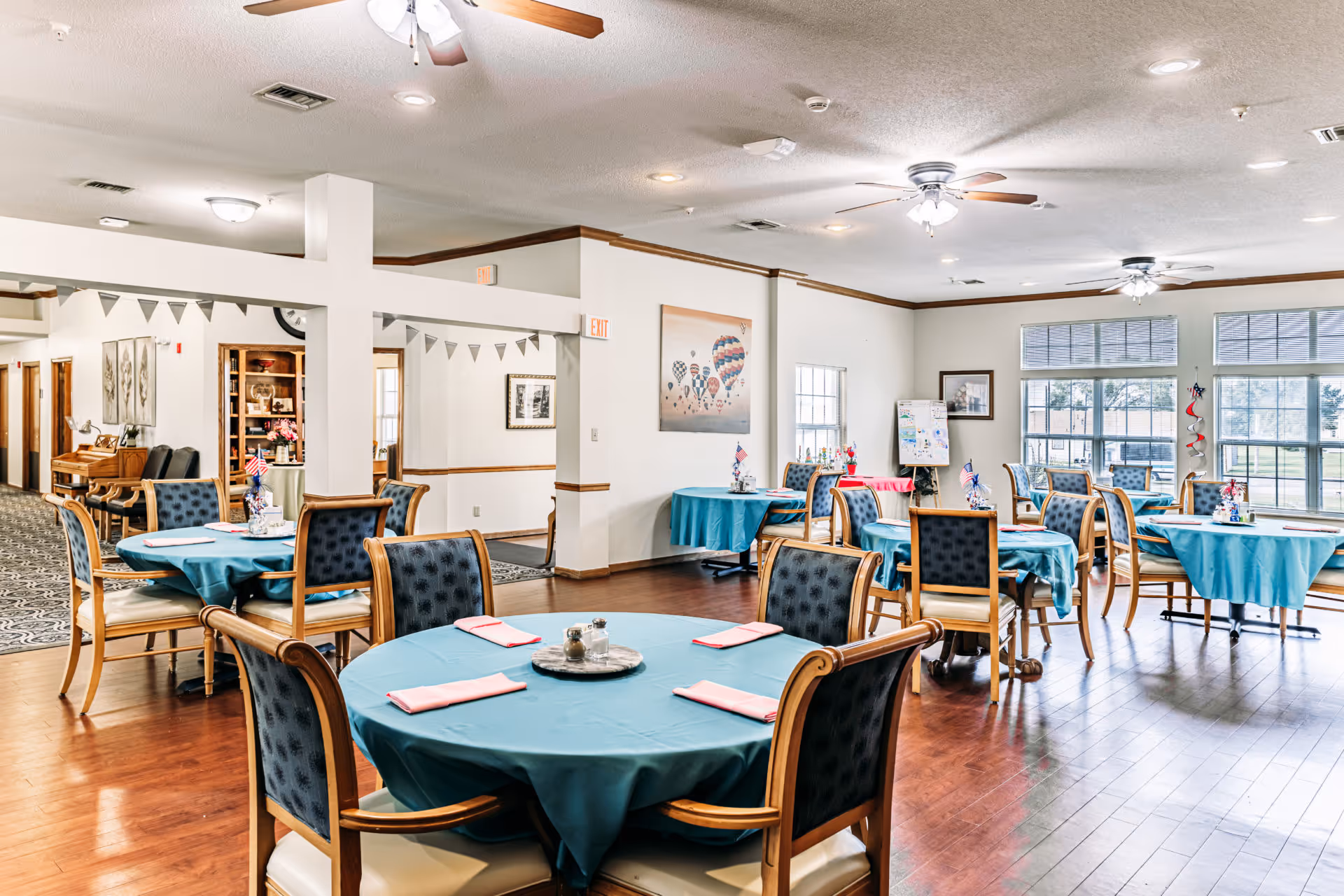 A bright dining room with multiple round tables covered with teal tablecloths and set with pink napkins. Each table is surrounded by wooden chairs with dark blue cushioned backs and cream seats. The room has large windows letting in natural light, ceiling fans with lights, and wooden flooring. Decorations include small American flags and patriotic-themed centerpieces on the tables. There is a painting of hot air balloons on one wall and an easel with drawings near the windows.