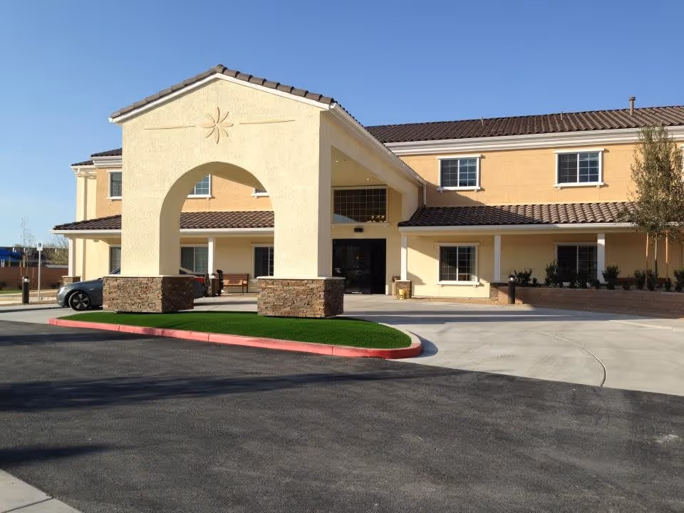 Exterior view of a senior living facility building with a beige and yellow facade, a tiled roof, and a large archway entrance supported by stone pillars. There is a driveway and a small landscaped area with green grass in front of the entrance under a clear blue sky.