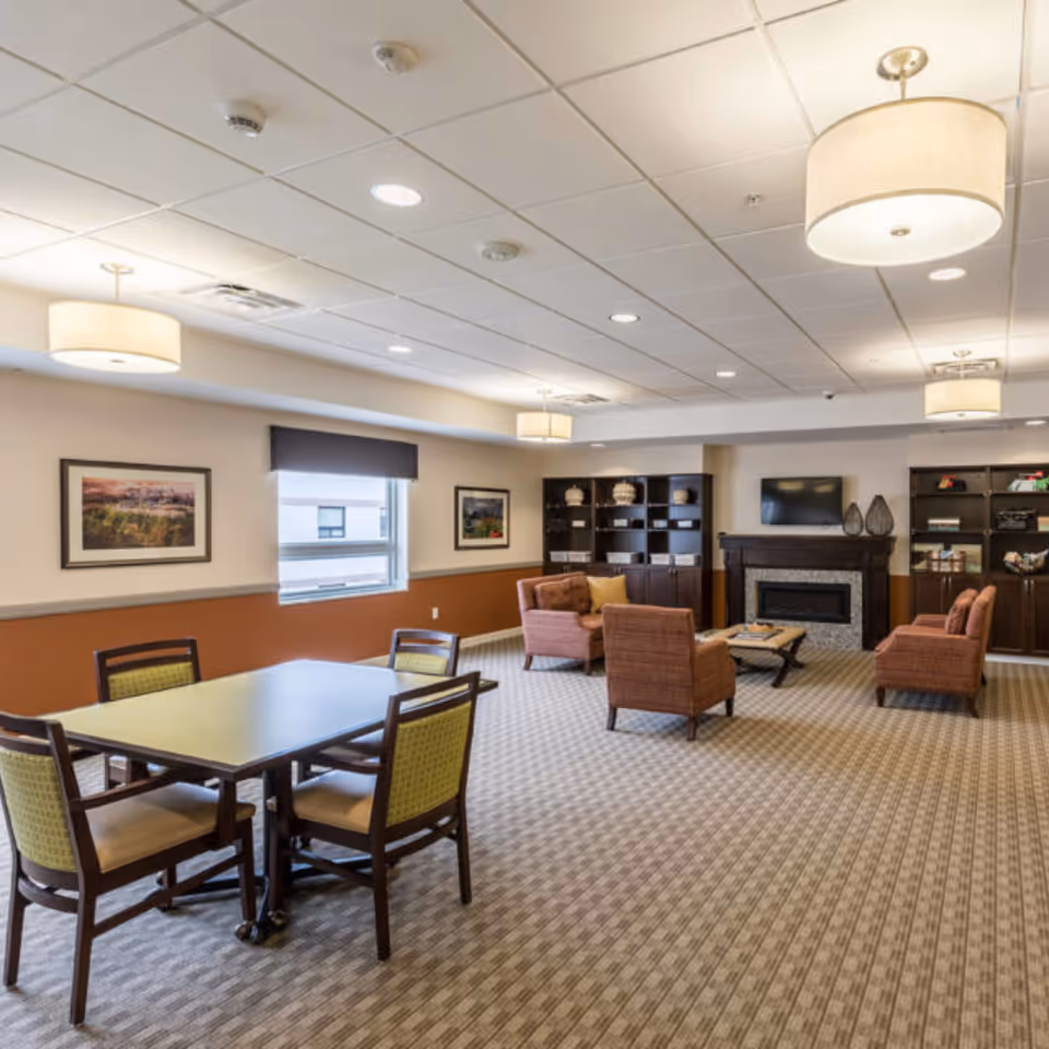 A spacious common area in a senior living facility featuring a table with four chairs in the foreground and a seating area with four armchairs around a coffee table near a fireplace. The room has carpeted flooring, built-in dark wood shelves with decorative items, framed pictures on the walls, and large ceiling lights providing warm illumination.